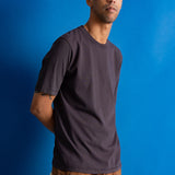 Man wearing dark brown t-shirt and dark amber cords against a bright blue backdrop.