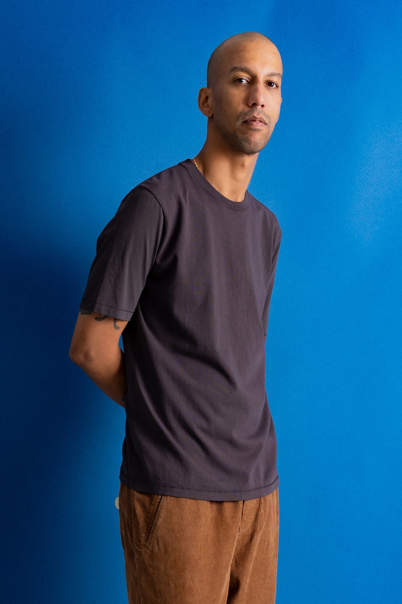Man wearing dark brown t-shirt and dark amber cords against a bright blue backdrop.