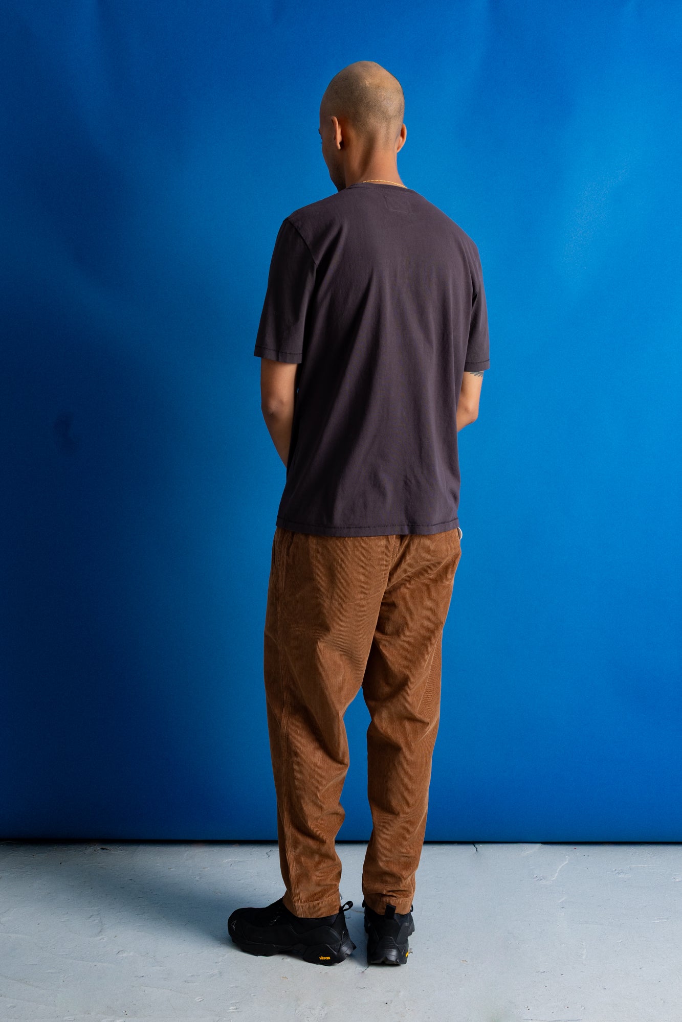 A man viewed from behind wearing a dark brown t-shirt and brown corduroy pants, standing against a vibrant blue studio backdrop.