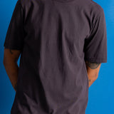 Man wearing dark brown t-shirt and dark amber cords against a bright blue backdrop.