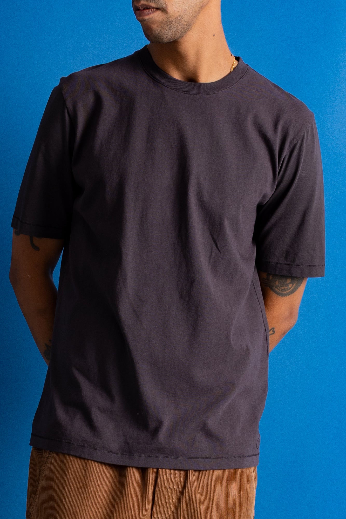 Man wearing dark brown t-shirt and dark amber cords against a bright blue backdrop.