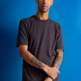 A man with a shaved head and tattoos wearing a dark charcoal gray t-shirt and brown pants, standing against a solid blue background.