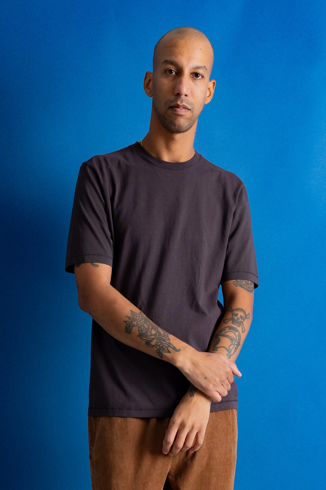 A man with a shaved head and tattoos wearing a dark charcoal gray t-shirt and brown pants, standing against a solid blue background.