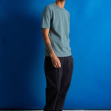 A man wearing a muted sage green crewneck t-shirt and black trousers, standing against a vibrant blue studio backdrop.