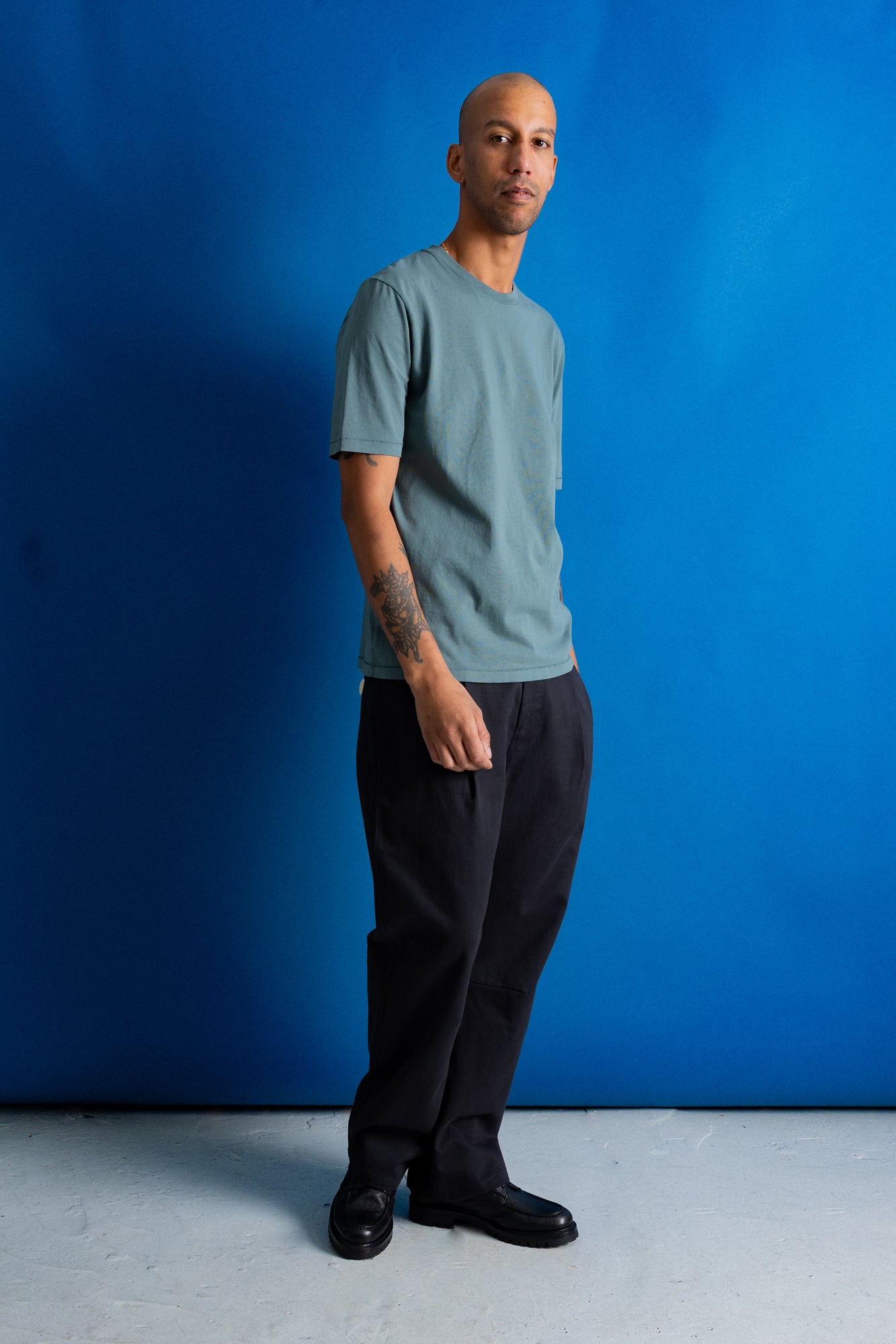 A man wearing a muted sage green crewneck t-shirt and black trousers, standing against a vibrant blue studio backdrop.