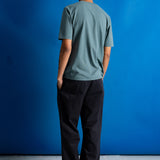 A man viewed from behind wearing a muted sage green t-shirt and black trousers, standing against a solid blue studio backdrop.