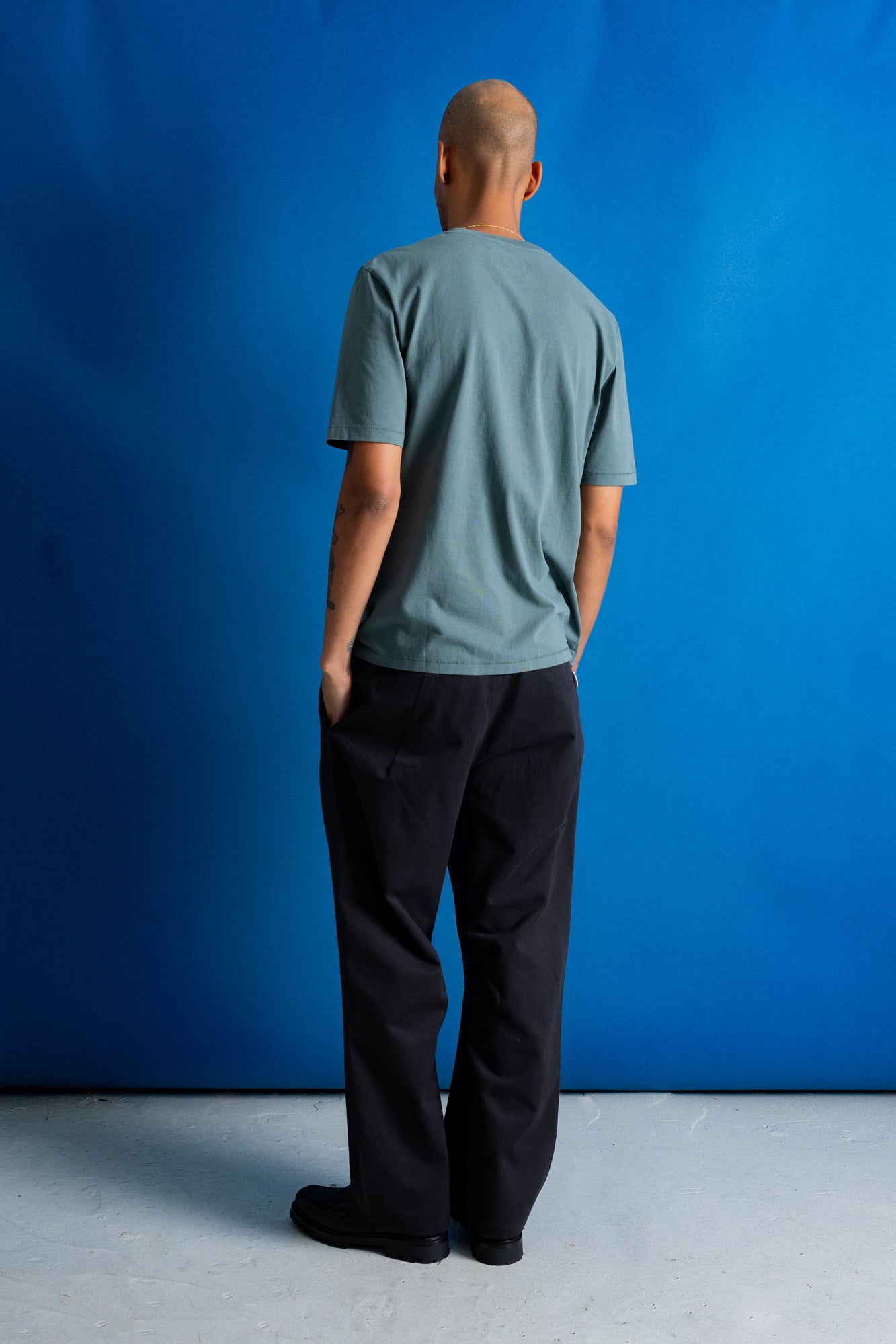 A man viewed from behind wearing a muted sage green t-shirt and black trousers, standing against a solid blue studio backdrop.