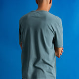 A man viewed from behind wearing a muted blue-green crewneck t-shirt and dark pants, standing against a solid blue backdrop.
