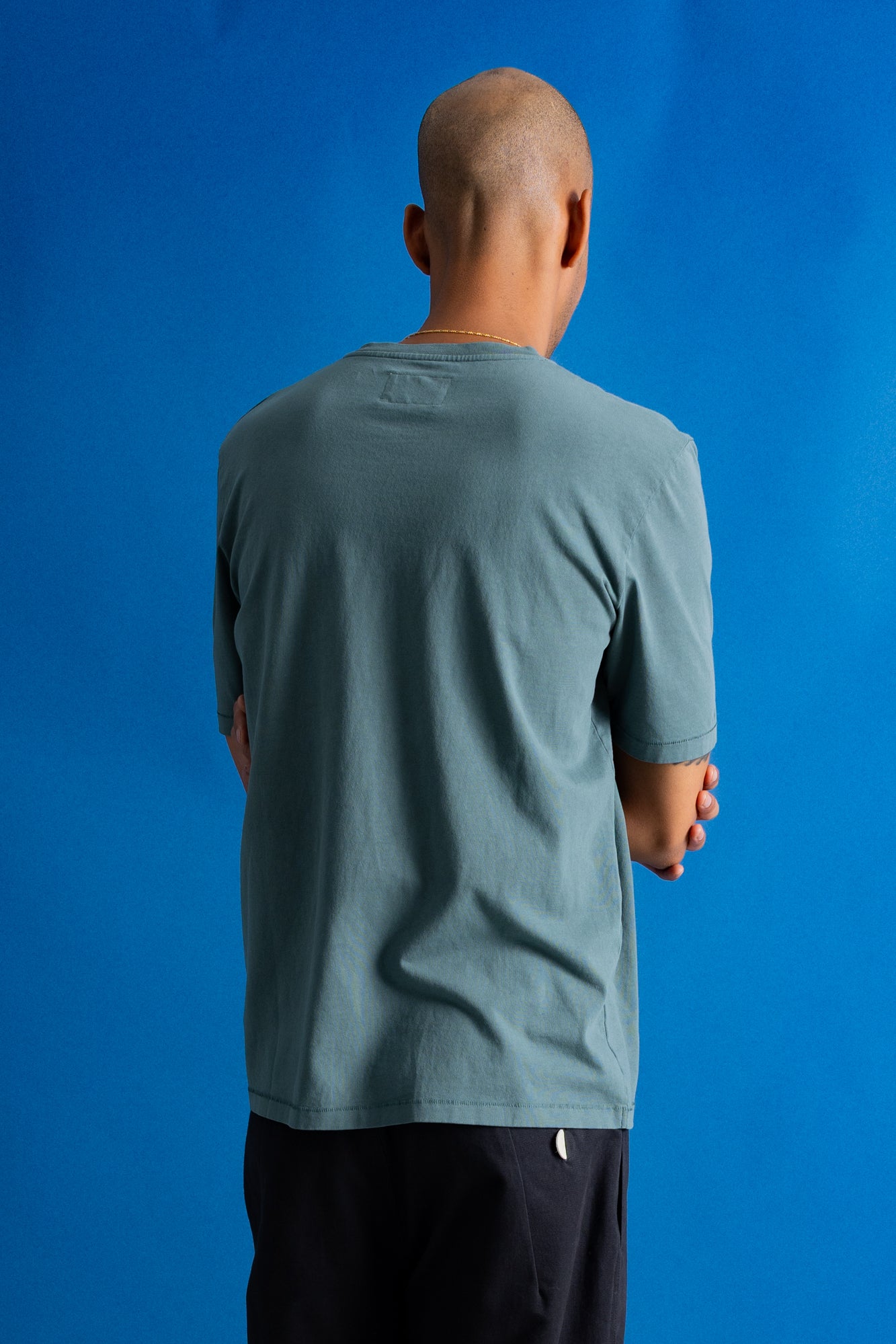 A man viewed from behind wearing a muted blue-green crewneck t-shirt and dark pants, standing against a solid blue backdrop.