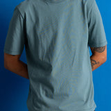 A man wearing a muted blue-green crewneck t-shirt with a subtle tonal topographic contour line pattern, set against a solid blue background.