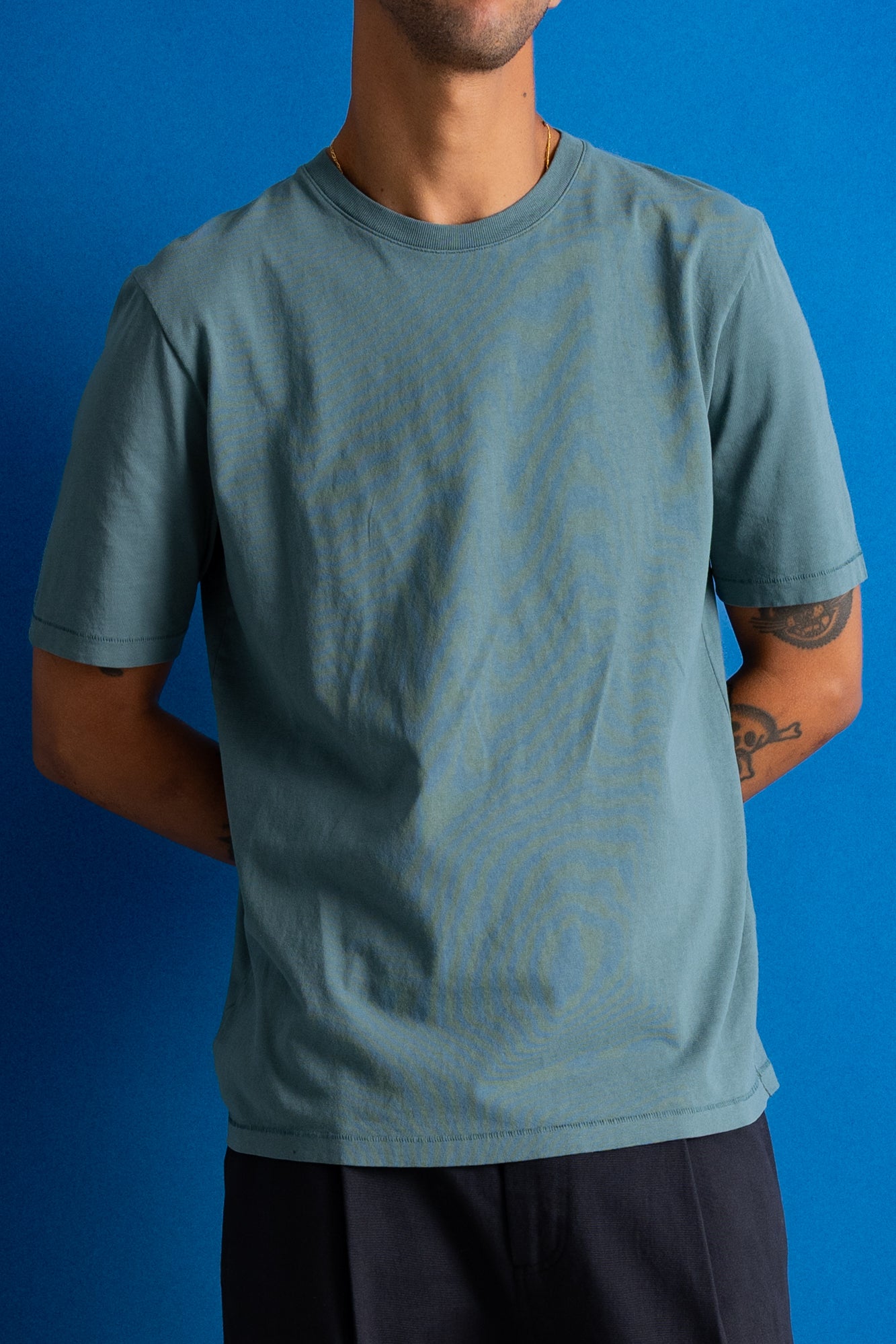 A man wearing a muted blue-green crewneck t-shirt with a subtle tonal topographic contour line pattern, set against a solid blue background.