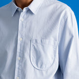 A close-up of a man wearing a light blue and white vertically striped button-down shirt with a chest pocket, set against a solid blue background.