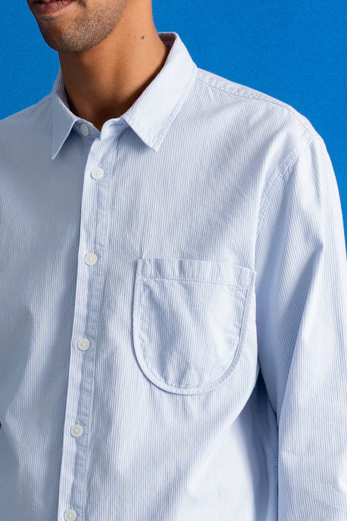 A close-up of a man wearing a light blue and white vertically striped button-down shirt with a chest pocket, set against a solid blue background.