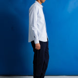 A man in a white long-sleeve button-down shirt, dark trousers, and black shoes stands in profile against a bright blue studio backdrop.