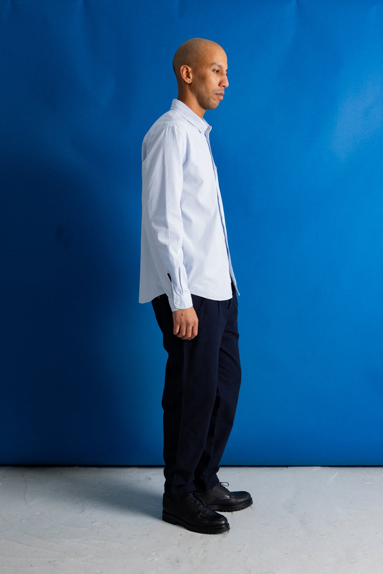A man in a white long-sleeve button-down shirt, dark trousers, and black shoes stands in profile against a bright blue studio backdrop.