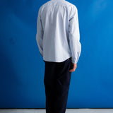 A man viewed from behind wearing a light blue long-sleeve button-up shirt, dark trousers, and black shoes, standing against a bright blue studio backdrop.