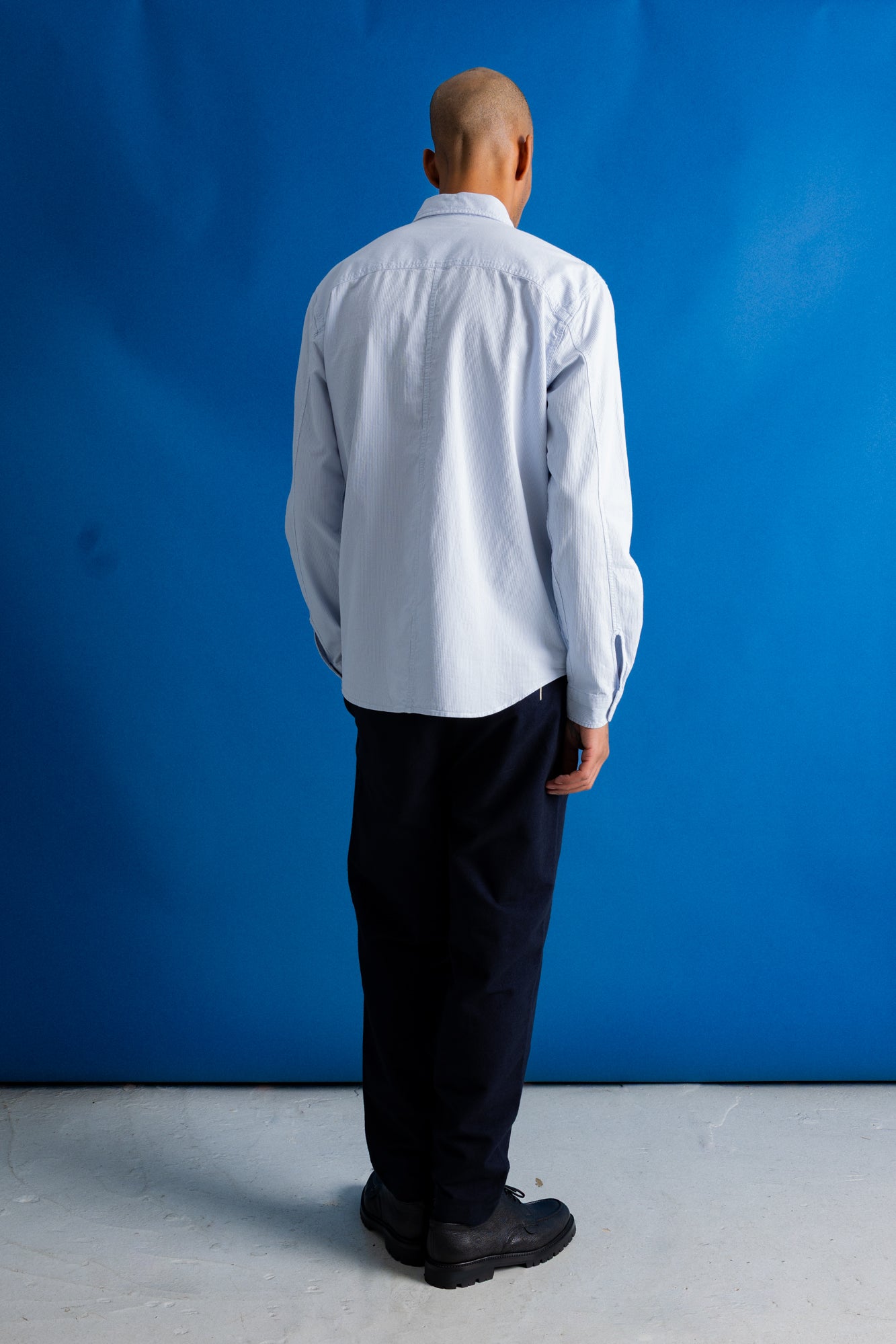 A man viewed from behind wearing a light blue long-sleeve button-up shirt, dark trousers, and black shoes, standing against a bright blue studio backdrop.