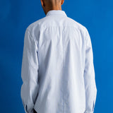 A man viewed from behind wearing a light blue long-sleeve button-down shirt and dark trousers, standing against a solid blue studio background.