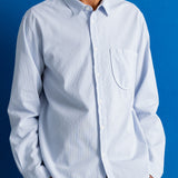 A man wearing a light blue long-sleeve button-down shirt with a single chest pocket, paired with dark trousers, set against a solid blue background.