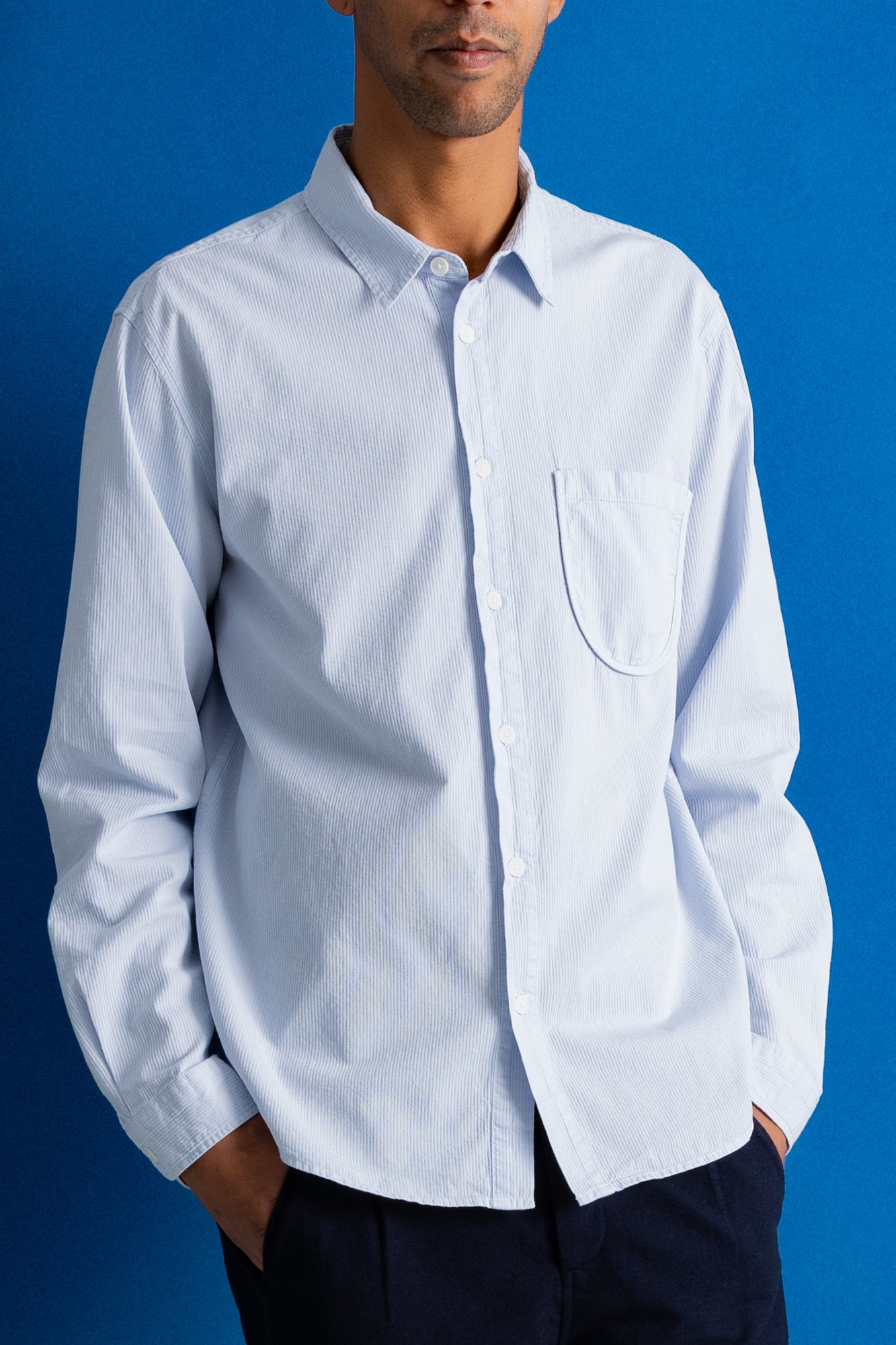 A man wearing a light blue long-sleeve button-down shirt with a single chest pocket, paired with dark trousers, set against a solid blue background.