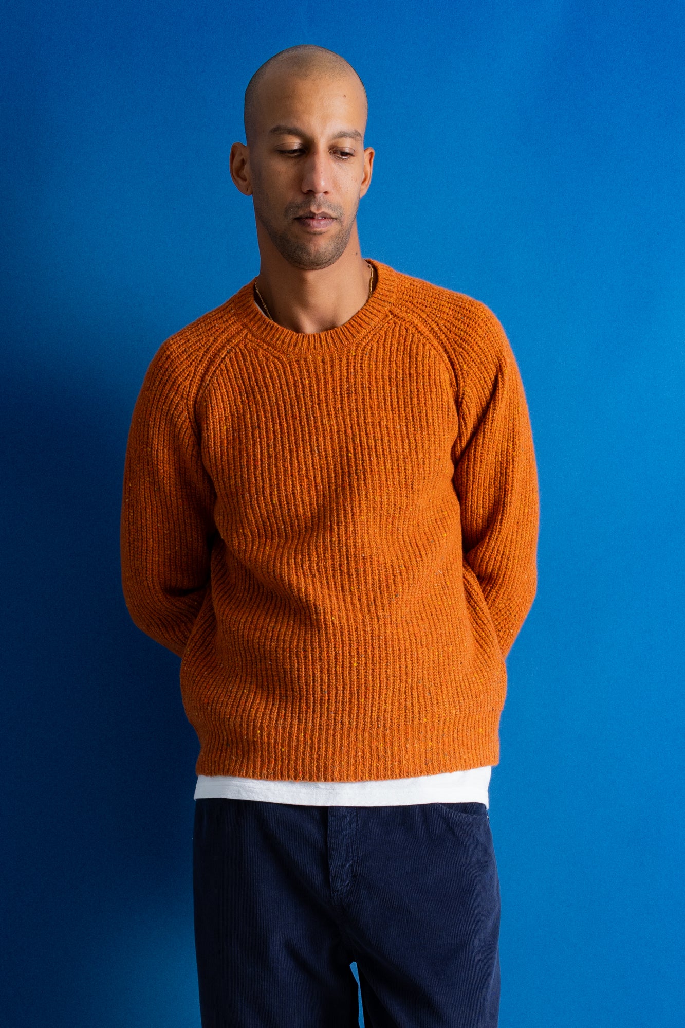 man wearing navy cord trousers, white t-shirt and orange sweater standing against a bright blue background