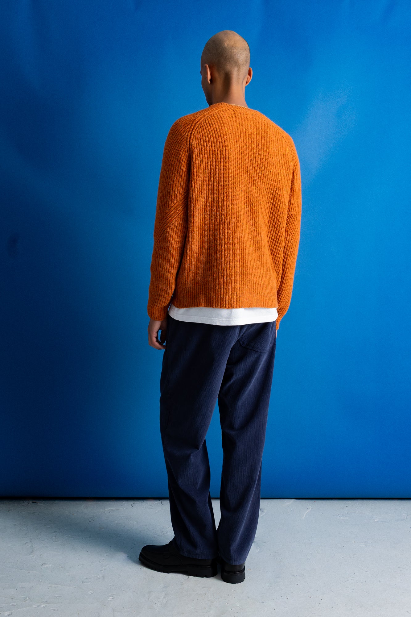 man wearing navy cord trousers, white t-shirt and orange sweater standing against a bright blue background