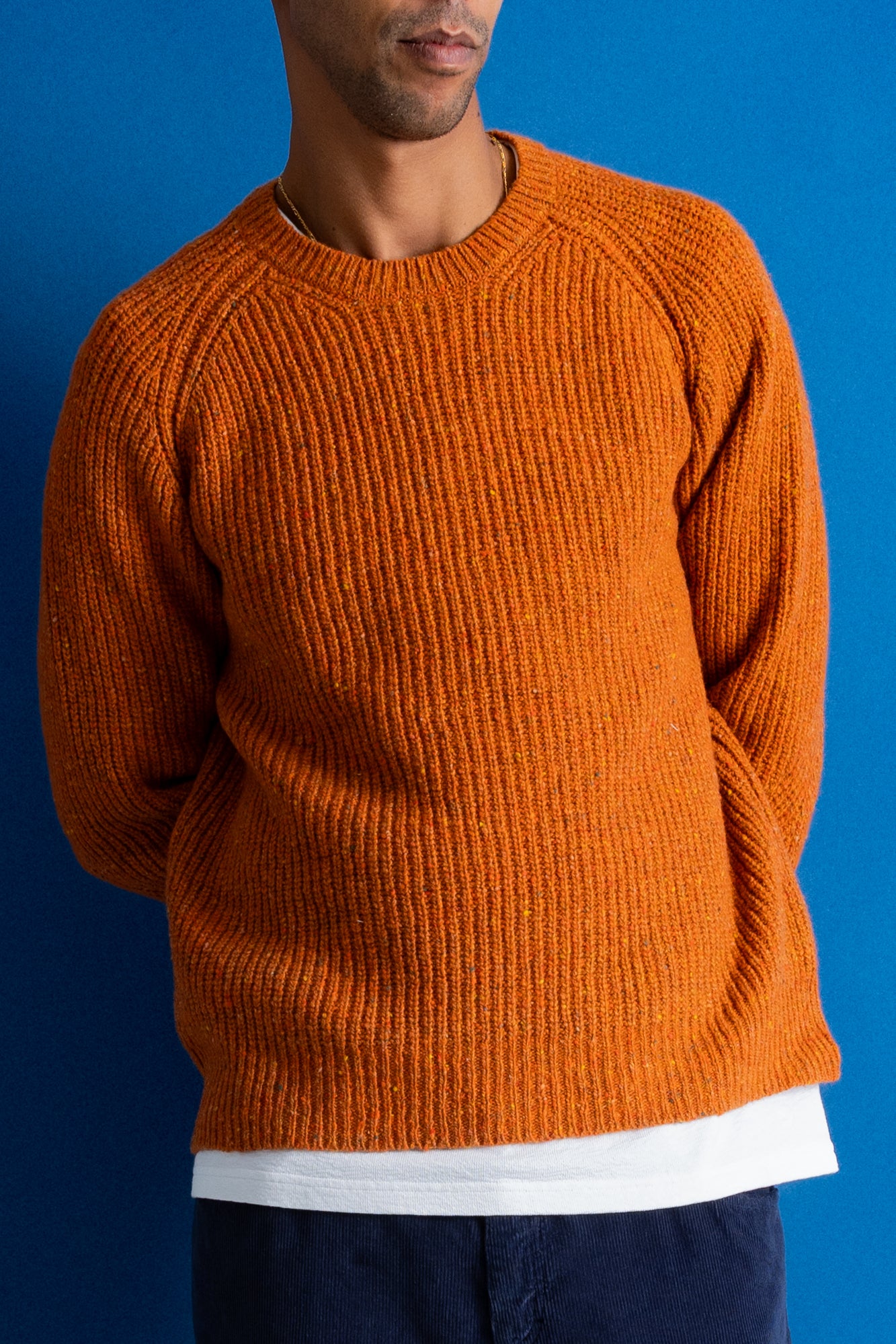 man wearing navy cord trousers, white t-shirt and orange sweater standing against a bright blue background