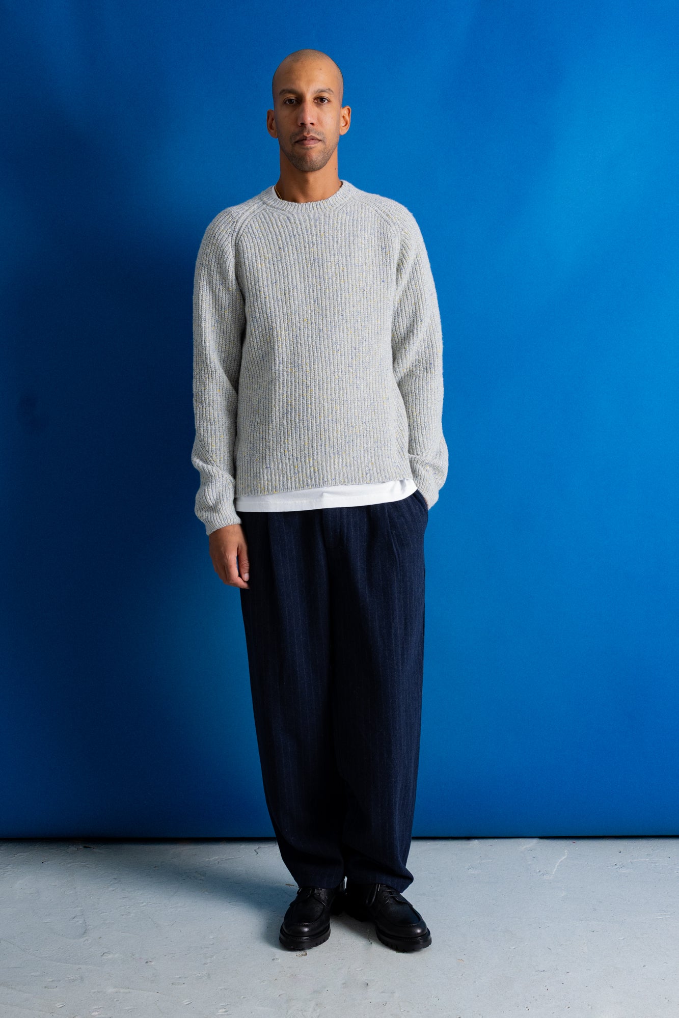 A man stands against a bright blue studio backdrop wearing a light gray ribbed knit sweater over a white undershirt, loose dark navy pants, and black shoes.