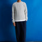 A man stands against a bright blue studio backdrop wearing a light gray ribbed knit sweater over a white undershirt, loose dark navy pants, and black shoes.