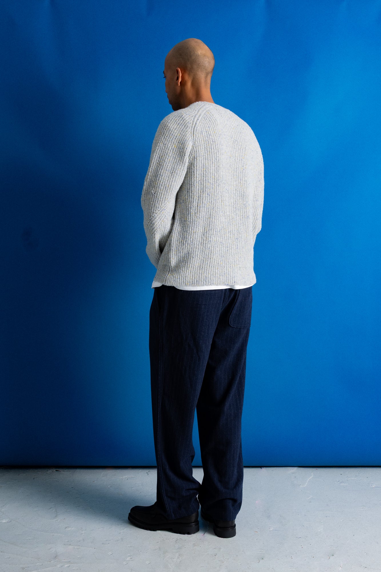 A man stands with his back to the camera wearing a light gray ribbed knit sweater and dark navy trousers, set against a solid blue studio backdrop.