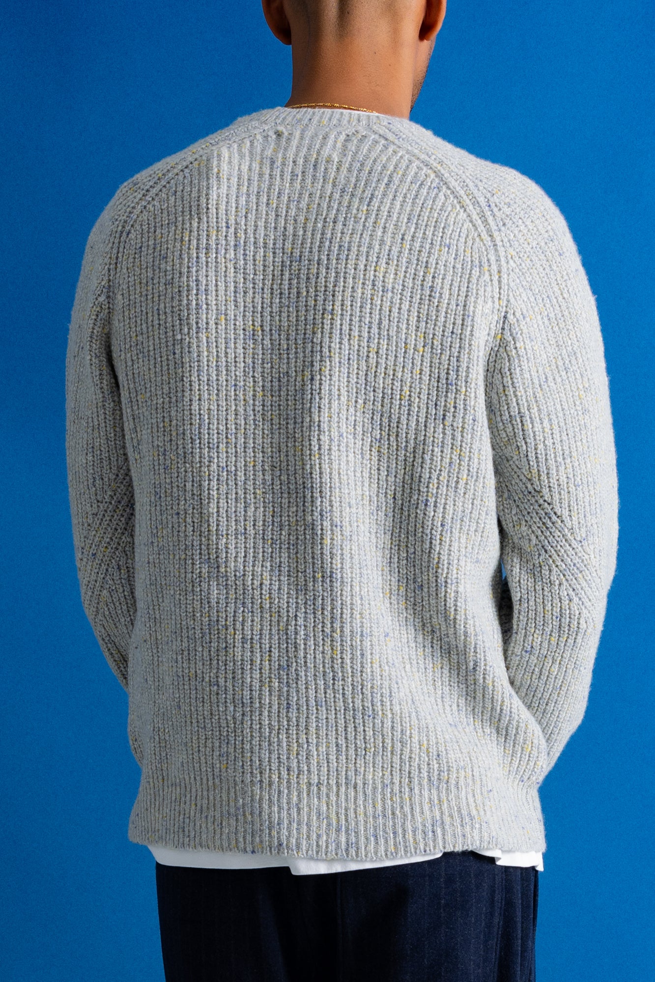A person shown from the back wearing a light gray ribbed knit sweater over a white shirt, against a solid blue background.