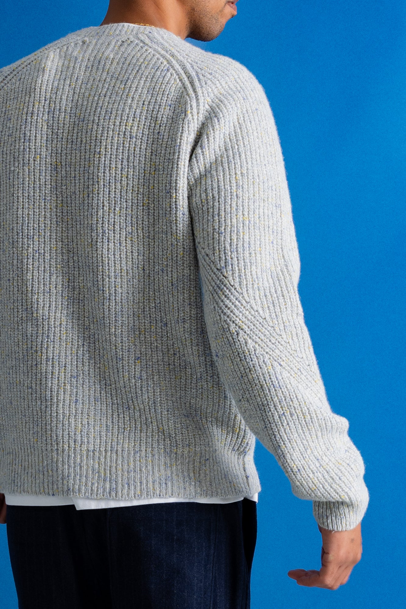 A man wearing a light gray ribbed knit sweater, shown from the back against a solid blue background.