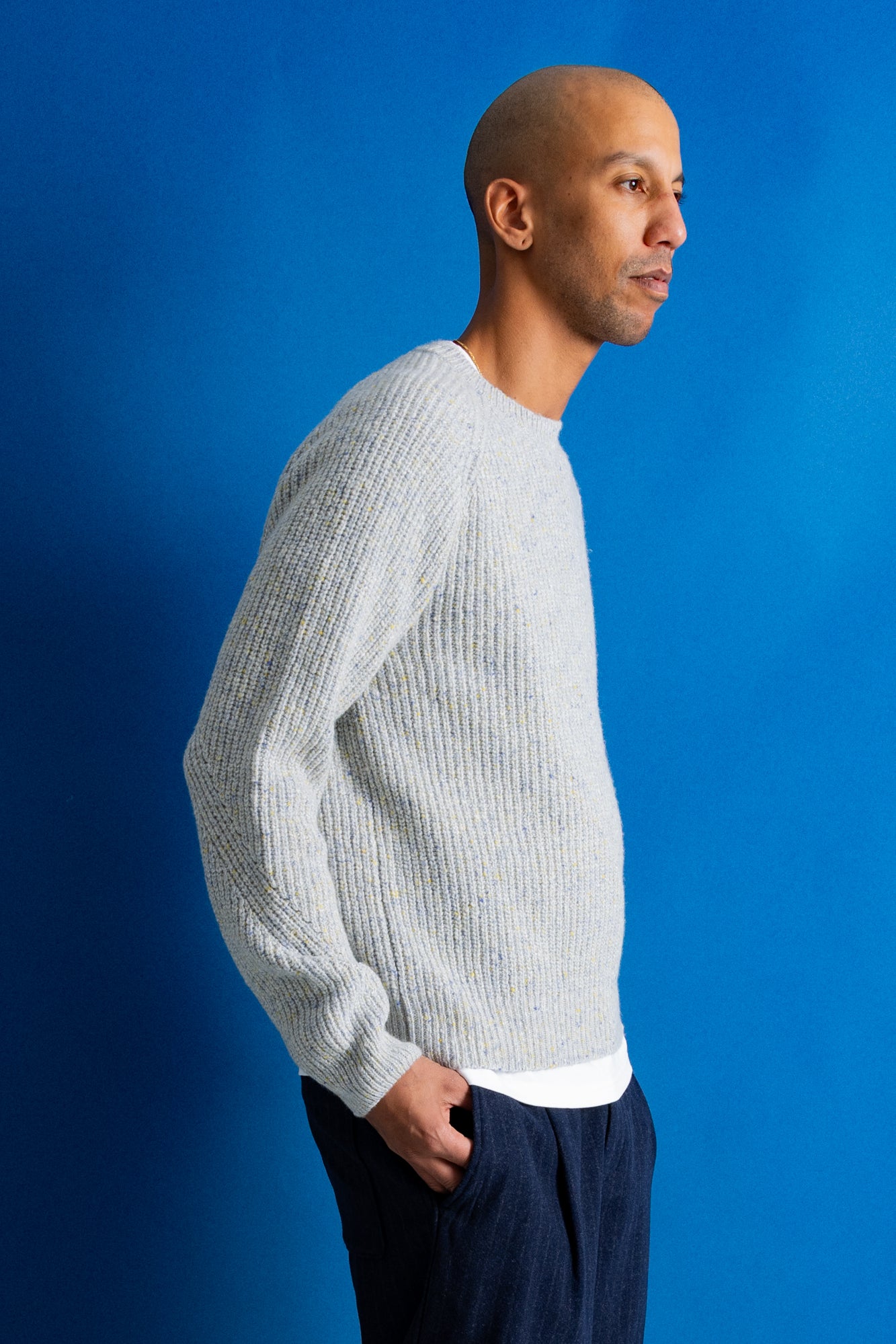 A man in profile wearing a light gray ribbed knit sweater and dark navy pants, standing against a solid blue background.