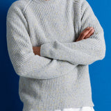 A man wearing a light gray ribbed knit sweater with subtle yellow and blue flecks, paired with a white undershirt and dark pants, standing against a solid blue background.