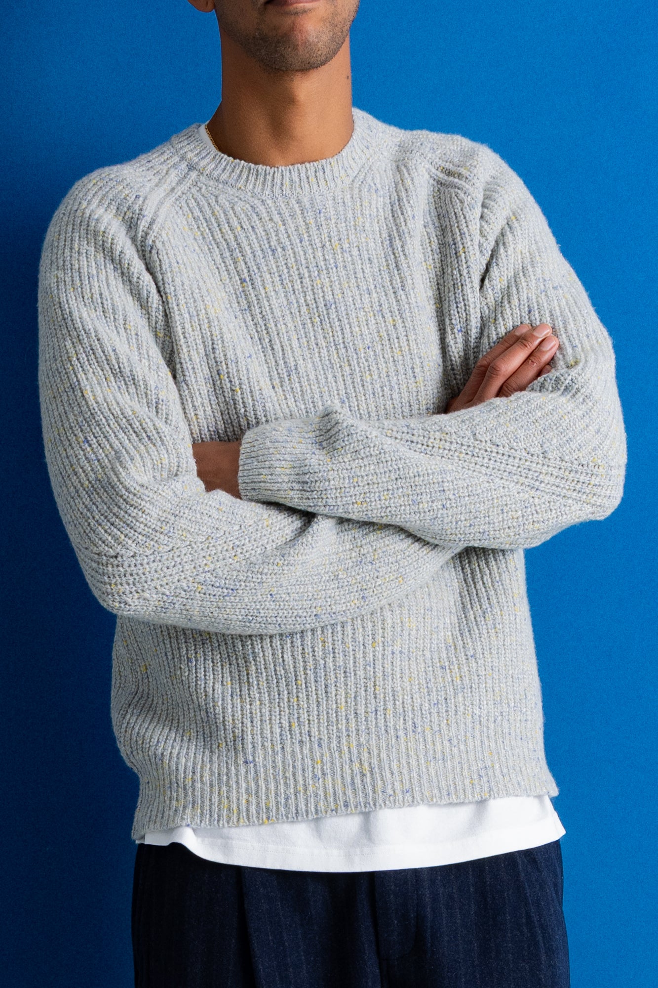A man wearing a light gray ribbed knit sweater with subtle yellow and blue flecks, paired with a white undershirt and dark pants, standing against a solid blue background.