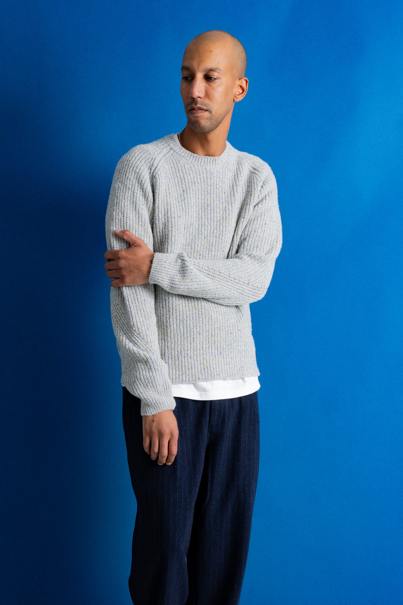A man wearing a light gray ribbed knit sweater over a white undershirt and dark navy pants, standing against a solid blue background.