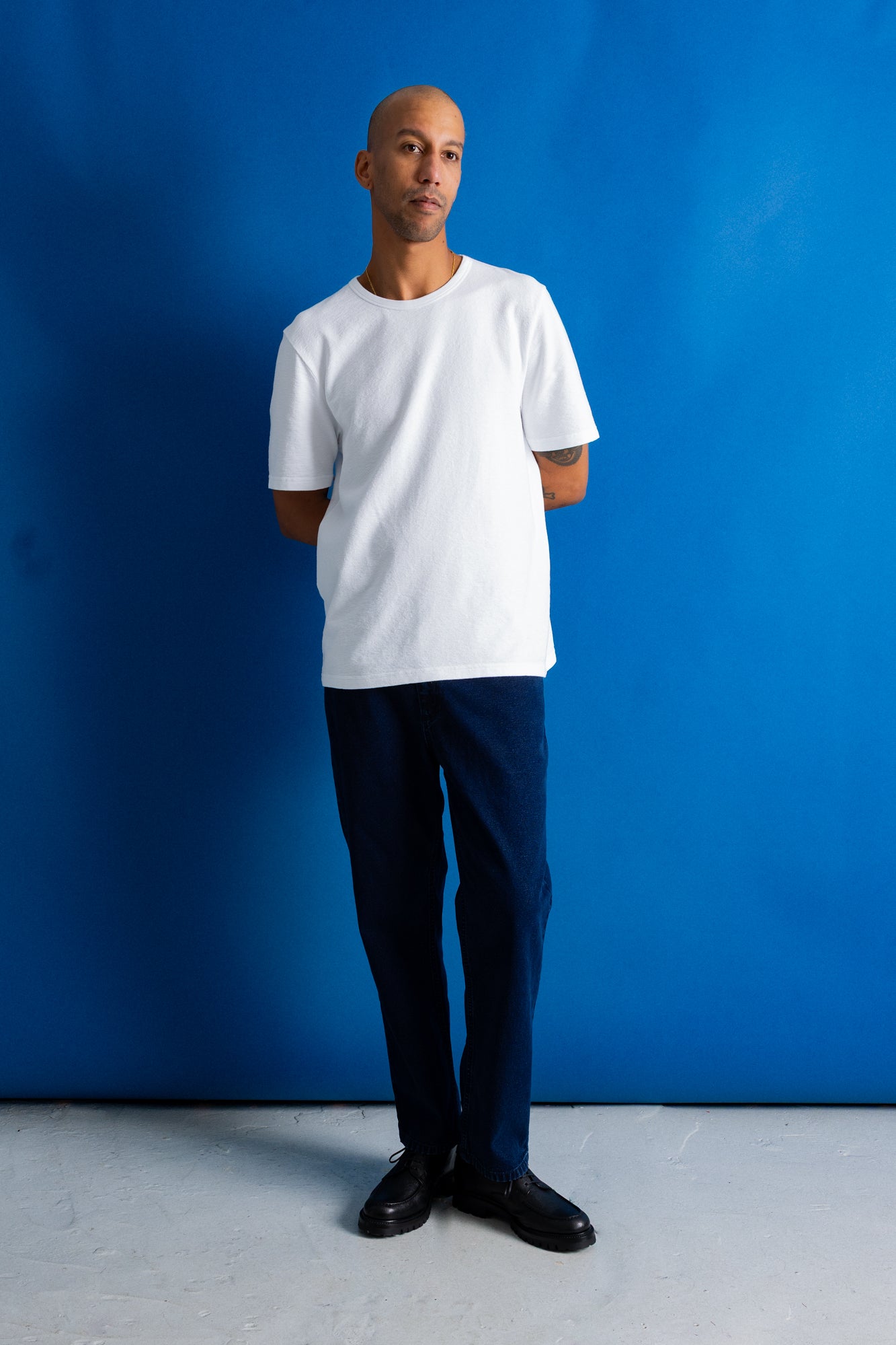A man wearing a plain white crew-neck t-shirt, dark navy trousers, and black shoes, standing against a vibrant blue studio backdrop.