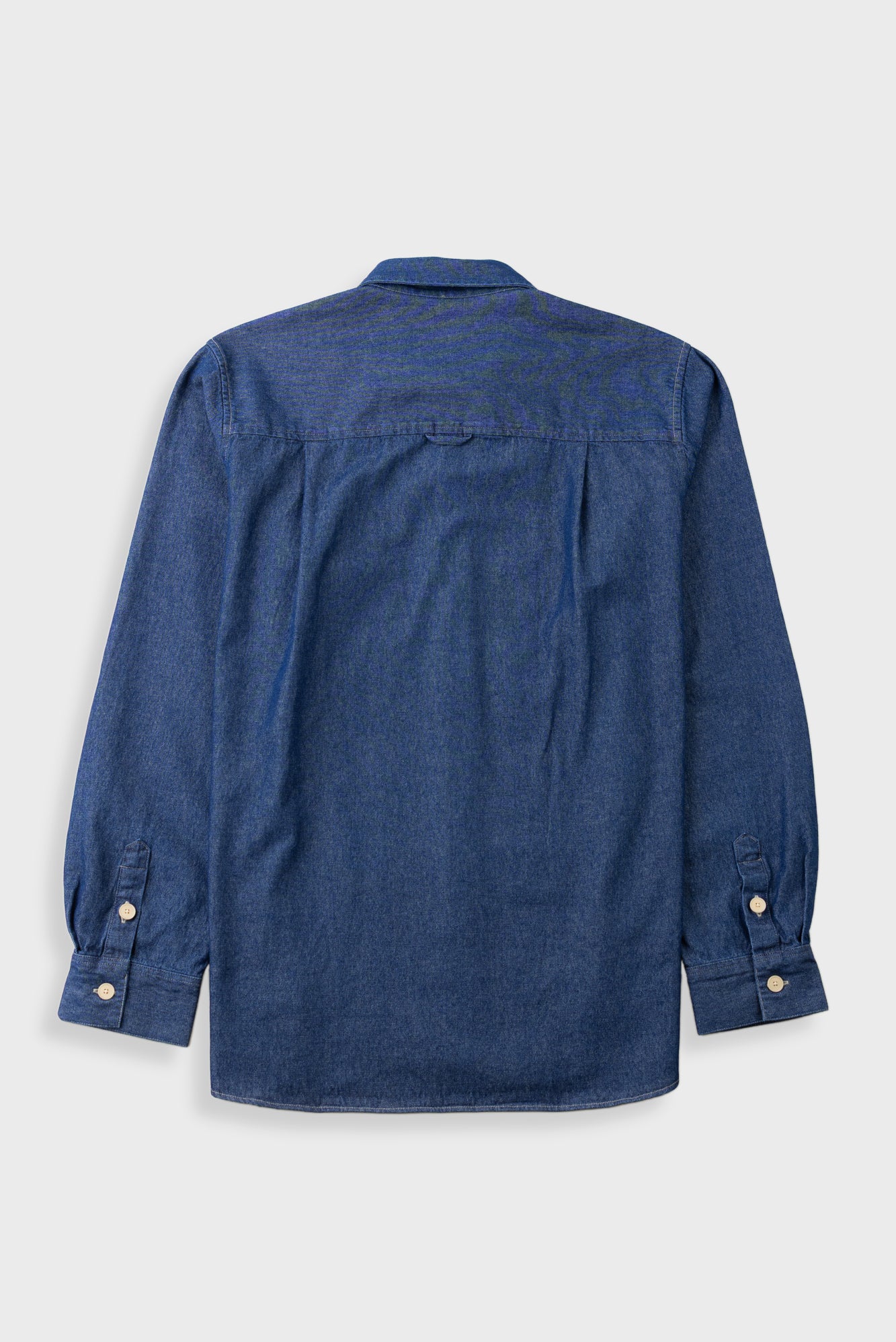 A blue denim long-sleeve button-up shirt shown from the back, featuring a collar, chest pocket, and buttoned cuffs, photographed on a light gray background.