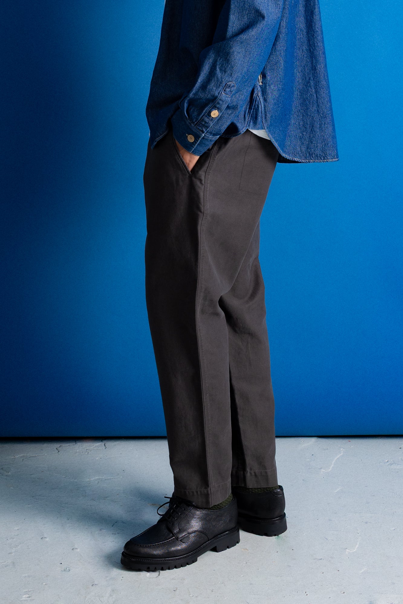 A person shown from the waist down wearing dark charcoal gray trousers, a blue denim shirt, and black leather shoes, standing against a vibrant blue studio backdrop.