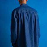 A man viewed from behind wearing a blue denim button-up shirt and dark trousers, standing against a solid blue studio background.