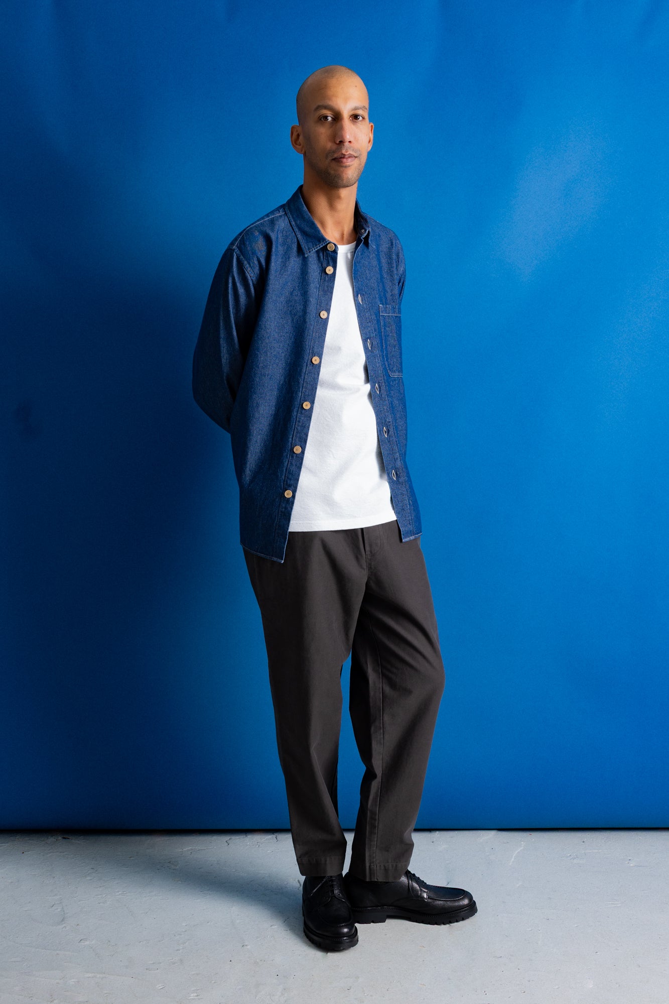 A man wearing a blue denim button-up shirt and dark trousers, standing against a solid blue studio background.