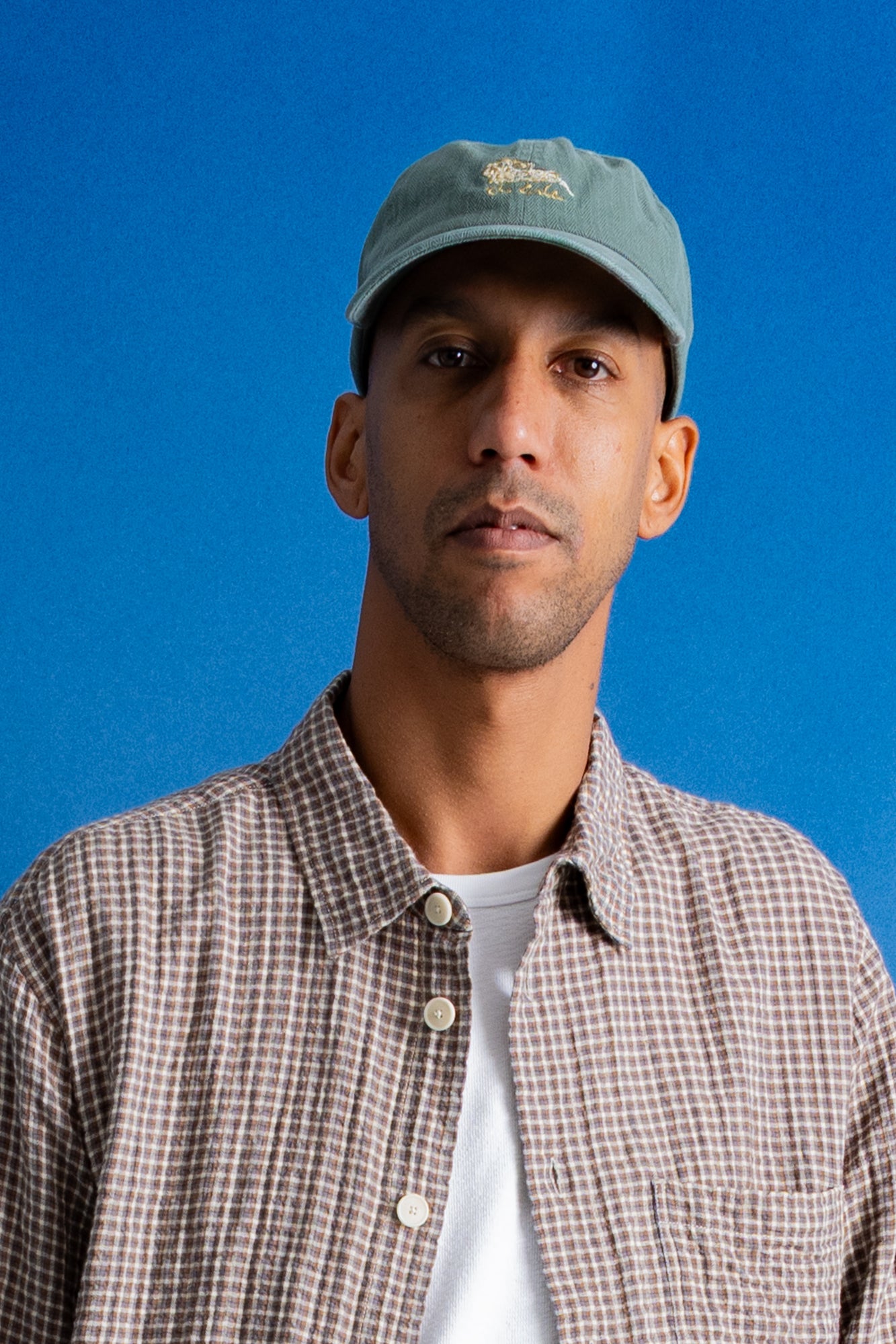 A man wearing a light blue baseball cap with a small embroidered logo, a brown and white checkered button-down shirt over a white t-shirt, set against a solid blue background.