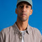 A man wearing a light blue baseball cap with a small embroidered logo, a brown and white checkered button-down shirt over a white t-shirt, set against a solid blue background.
