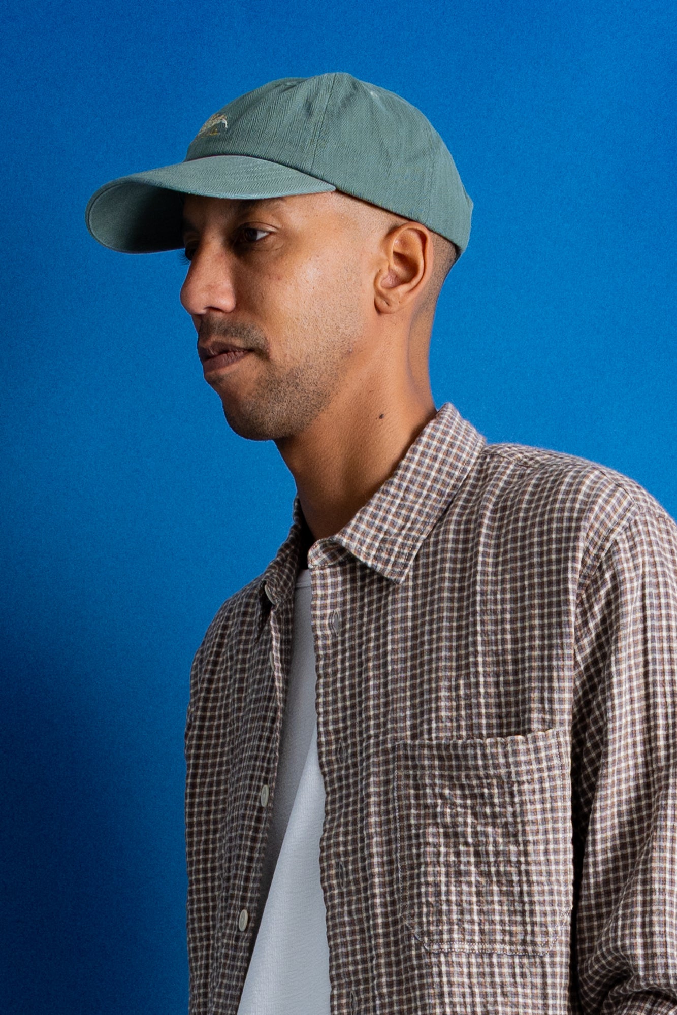 A man in profile wearing a muted sage green baseball cap, a brown and white checkered button-up shirt, and a white undershirt, set against a solid blue studio background.