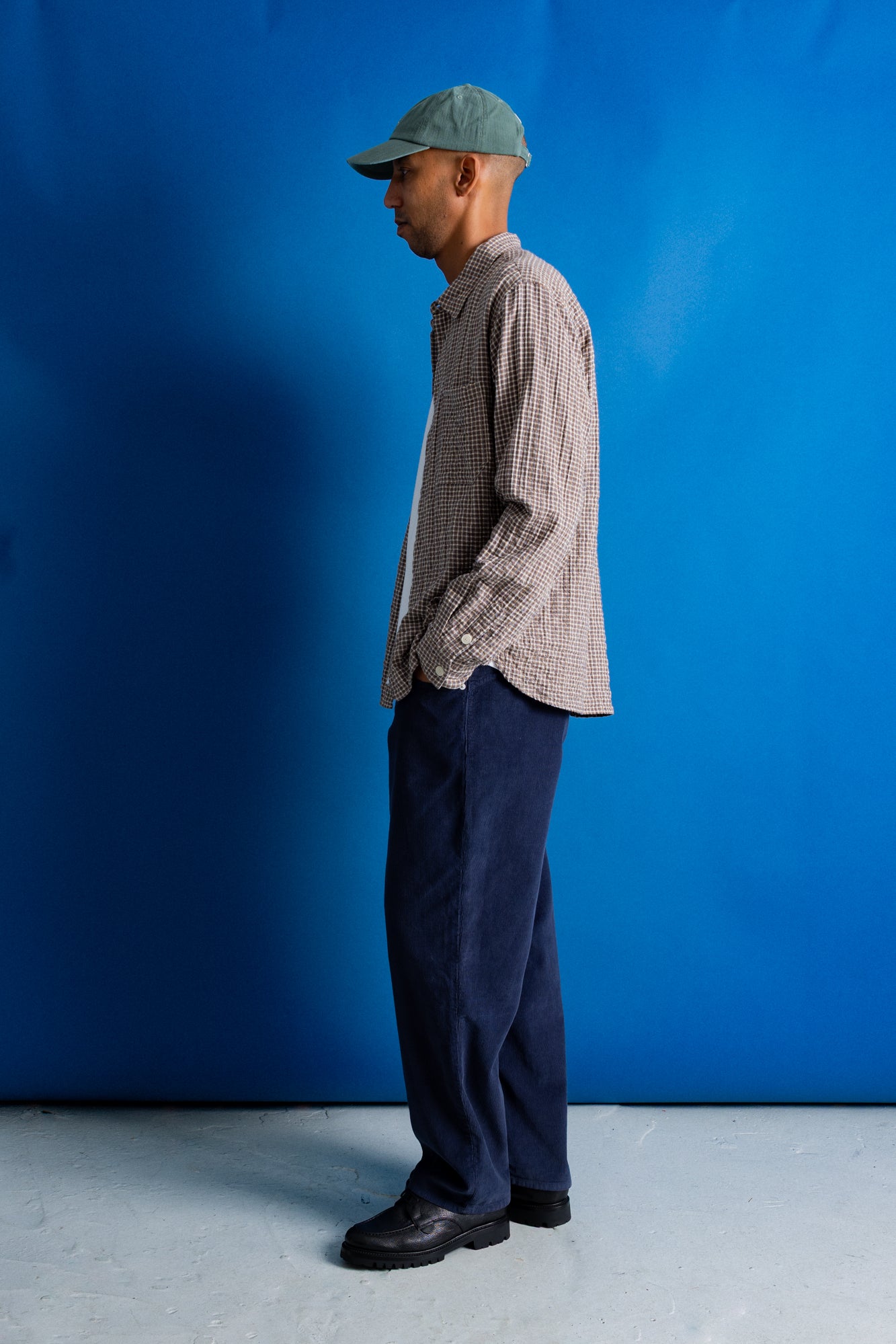 A man stands in profile wearing a green cap, a light brown and white checkered button-up shirt, dark navy trousers, and black shoes, set against a solid blue studio backdrop.