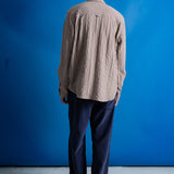 A man stands with his back to the camera wearing a beige and brown textured long-sleeve button-up shirt, navy blue trousers, and a green baseball cap against a bright blue studio backdrop.