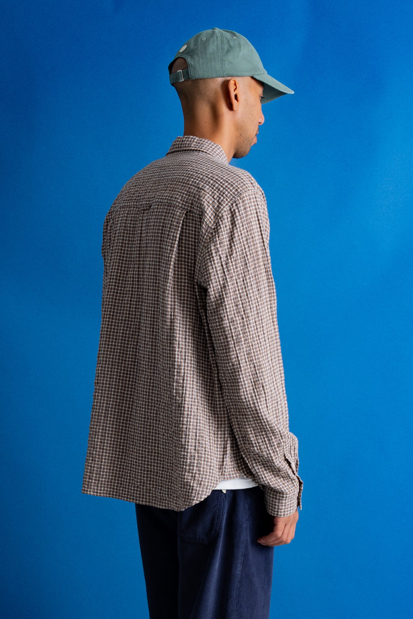A man wearing a brown and beige checkered long-sleeve shirt, navy pants, and a green cap, shown from a side-back angle against a solid blue background.