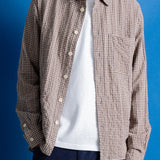 A man wearing a light brown and white checkered button-up shirt, dark navy trousers, and black shoes, set against a solid blue studio backdrop