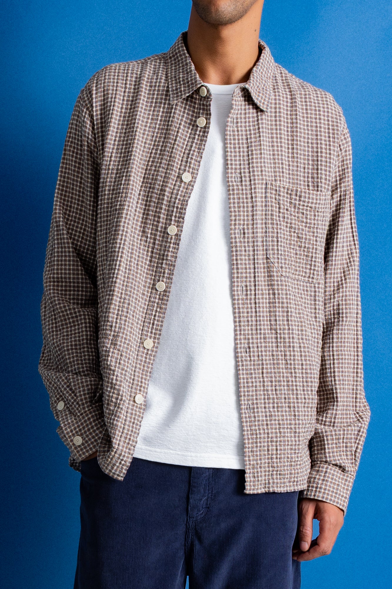 A man wearing a light brown and white checkered button-up shirt, dark navy trousers, and black shoes, set against a solid blue studio backdrop