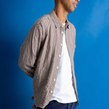A man wearing a brown and white checkered button-down shirt over a white t-shirt, navy pants, and a green cap, standing against a solid blue background.